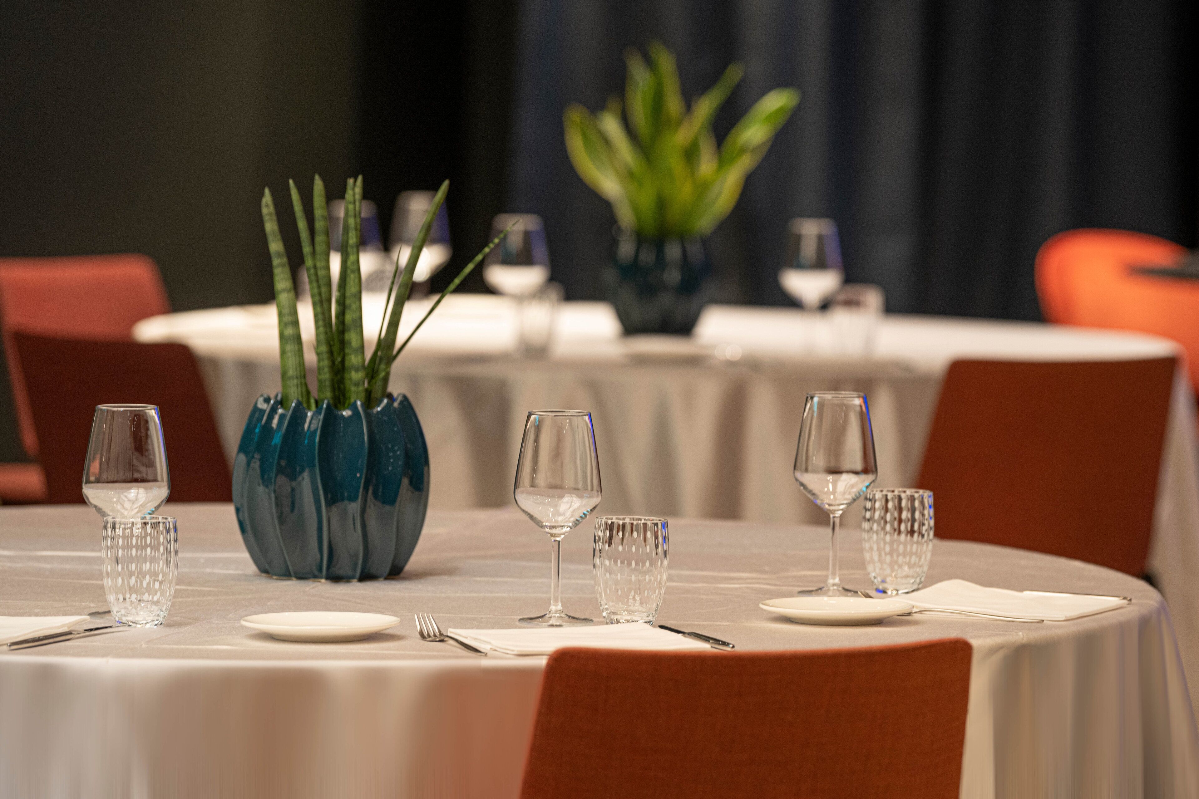 Une table à manger dressée avec des nappes blanches, de la vaisselle transparente et un vase bleu décoratif contenant des plantes vertes.