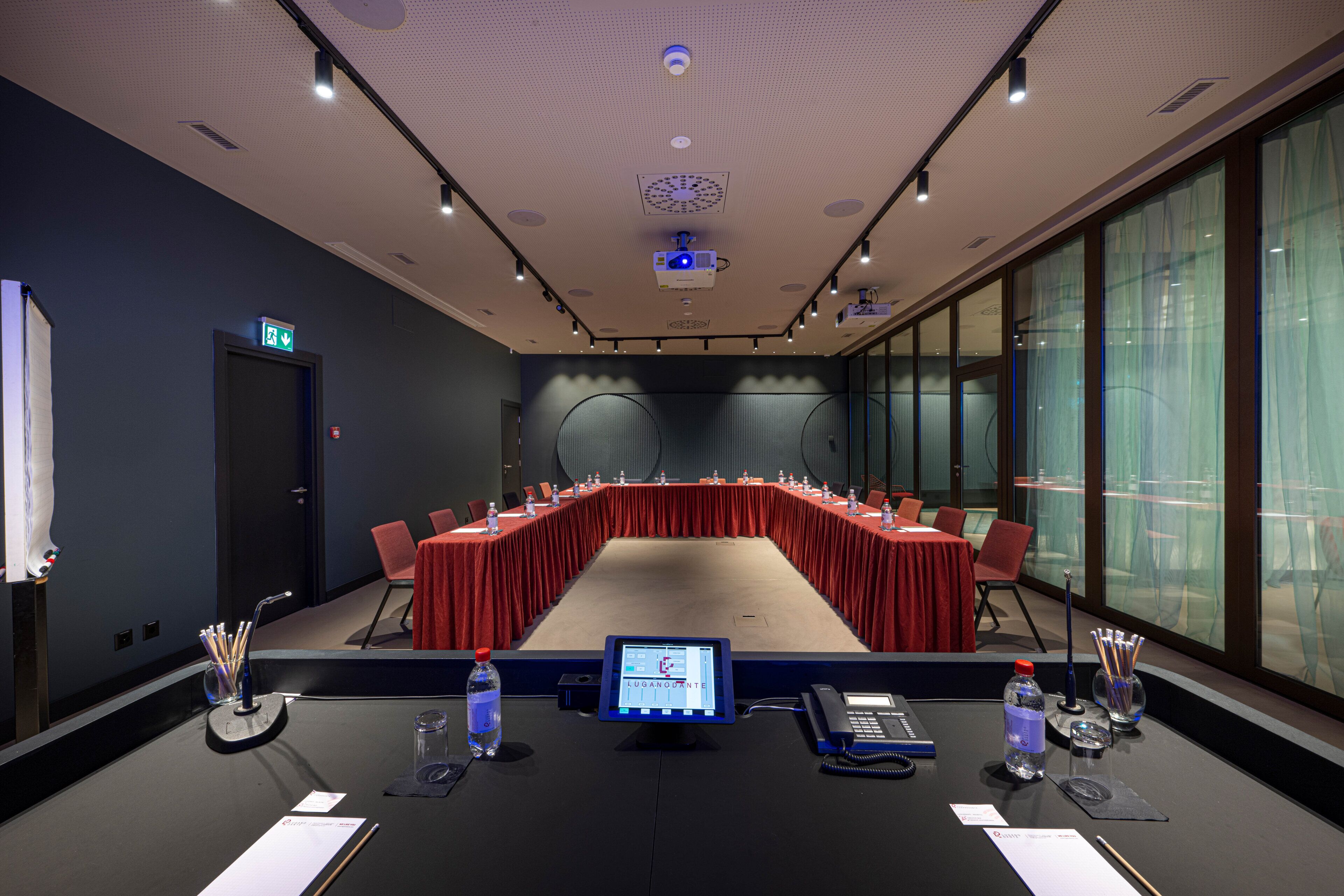 A conference room with a long table, chairs, a podium, a screen, and a water bottle.