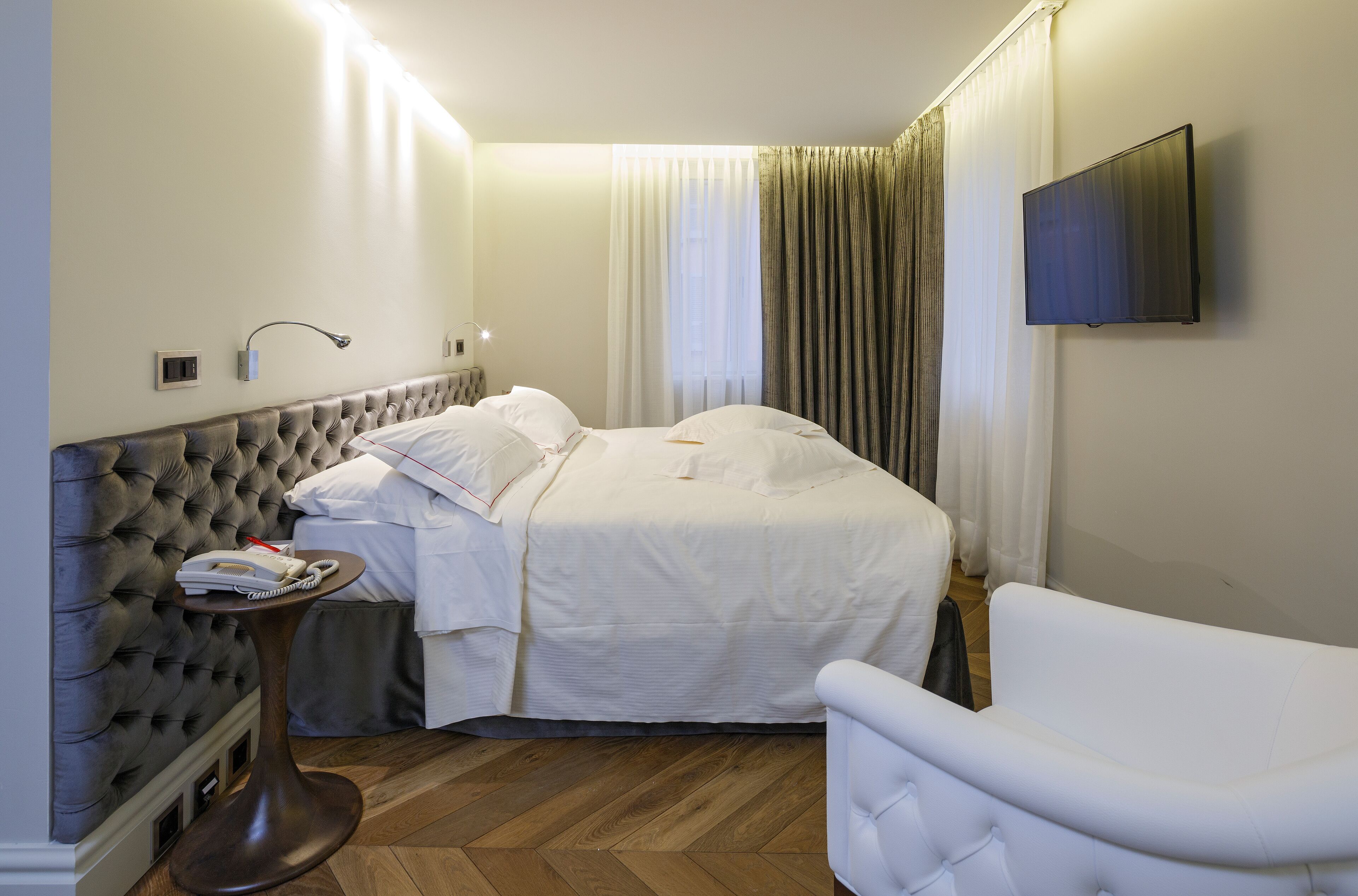 Una habitación de hotel con una cama, una mesita de noche con lámpara, un televisor montado en la pared y una silla.