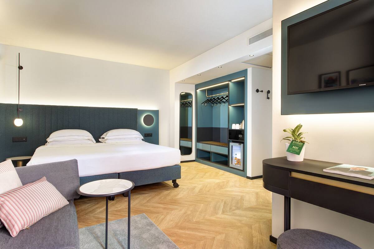 A modern hotel room with a bed, a sofa, a small table, a TV, and a wardrobe.