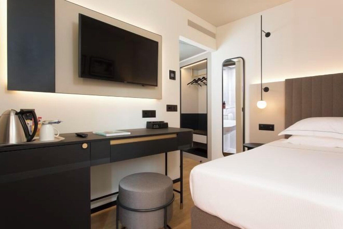 Una habitación de hotel moderna con una cama, un escritorio con una silla, un televisor y un armario.