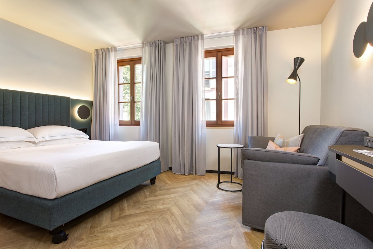 A modern hotel room with a large bed, a grey sofa, a small table, and a desk with a lamp.