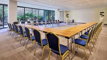 Un salón de conferencias con una larga mesa y sillas, ventanas amplias y vistas a árboles.
