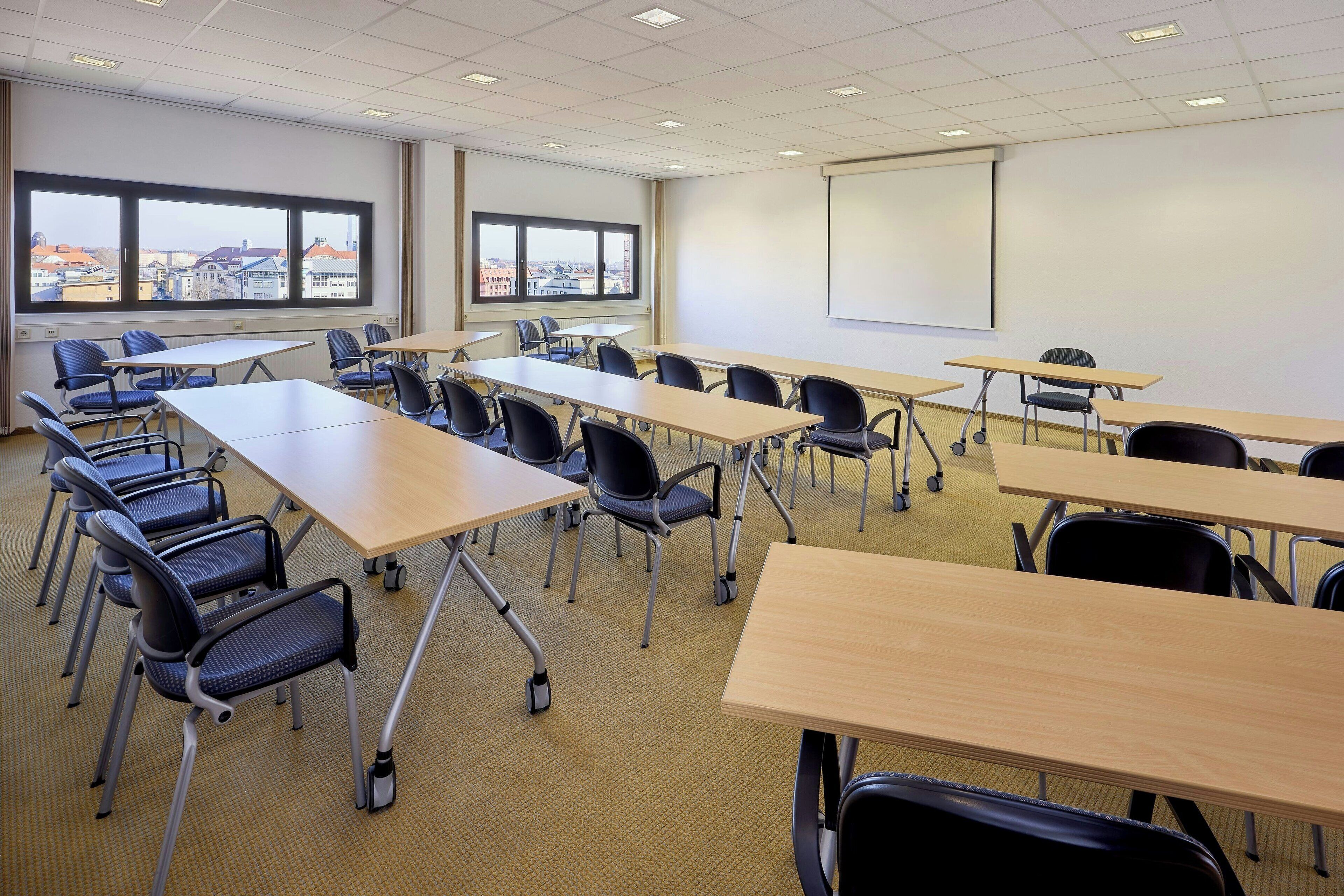 A classroom with rows of desks and chairs, large windows with a city view, and a projection screen on the wall.
