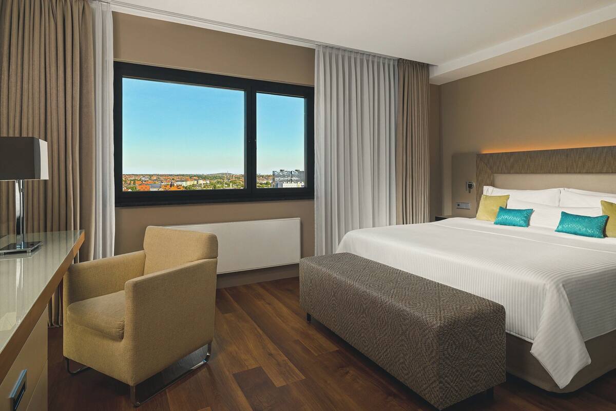 Habitación de hotel con una cama grande, un escritorio, una silla y vistas de la ciudad a través de un ventanal amplio.