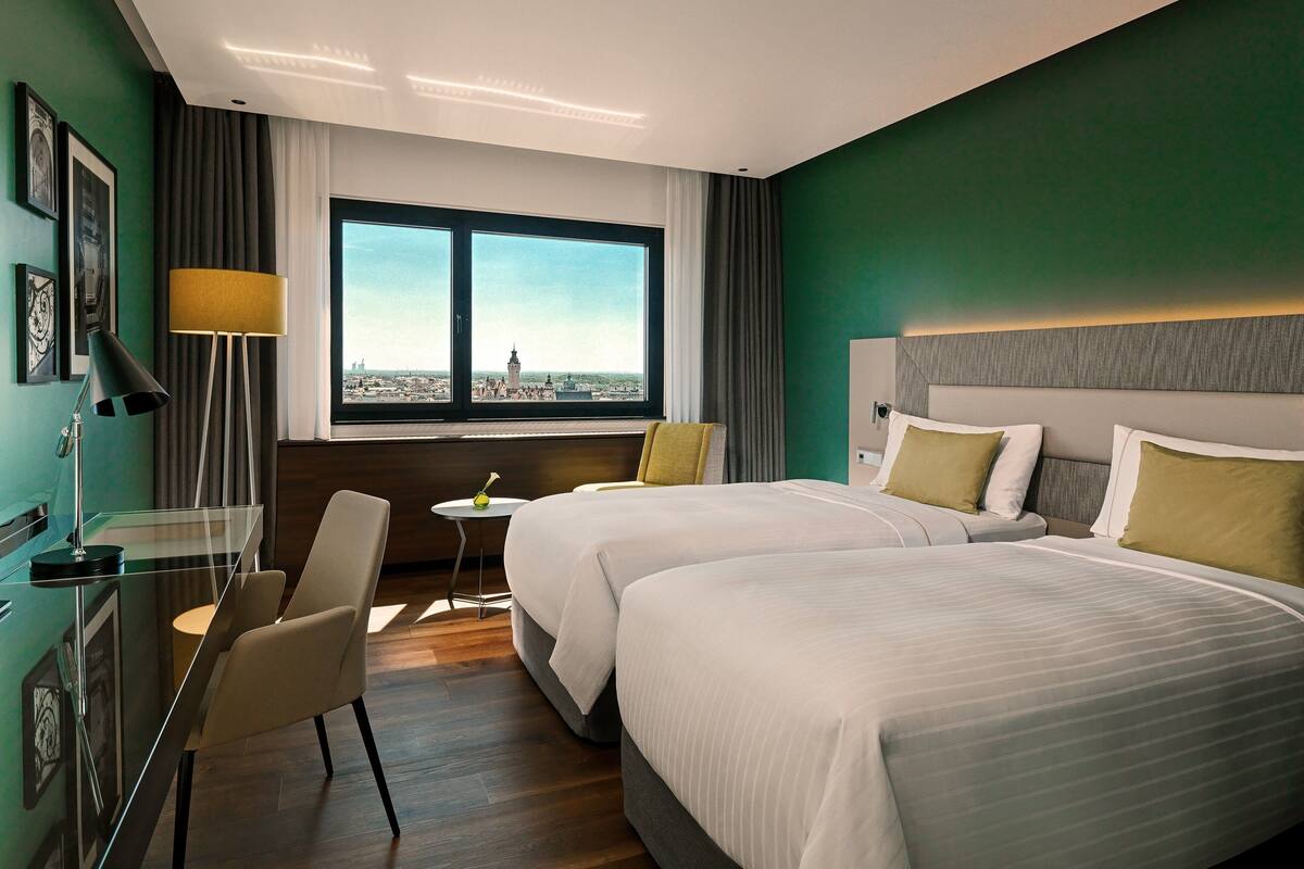 Habitación de hotel con dos camas, un escritorio, una silla, una lámpara y un ventanal con vistas a la ciudad.