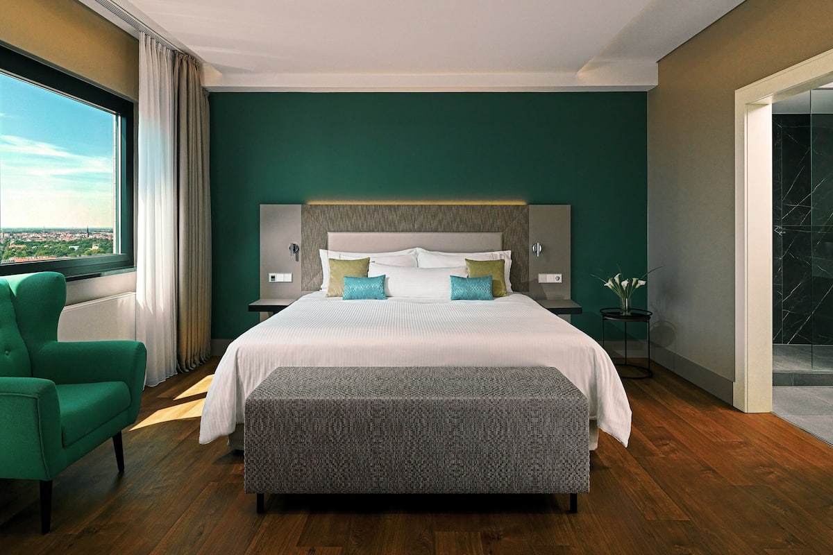 Una habitación de hotel moderna con una cama grande, un sillón verde, una mesita auxiliar y vistas de la ciudad a través de un ventanal amplio.
