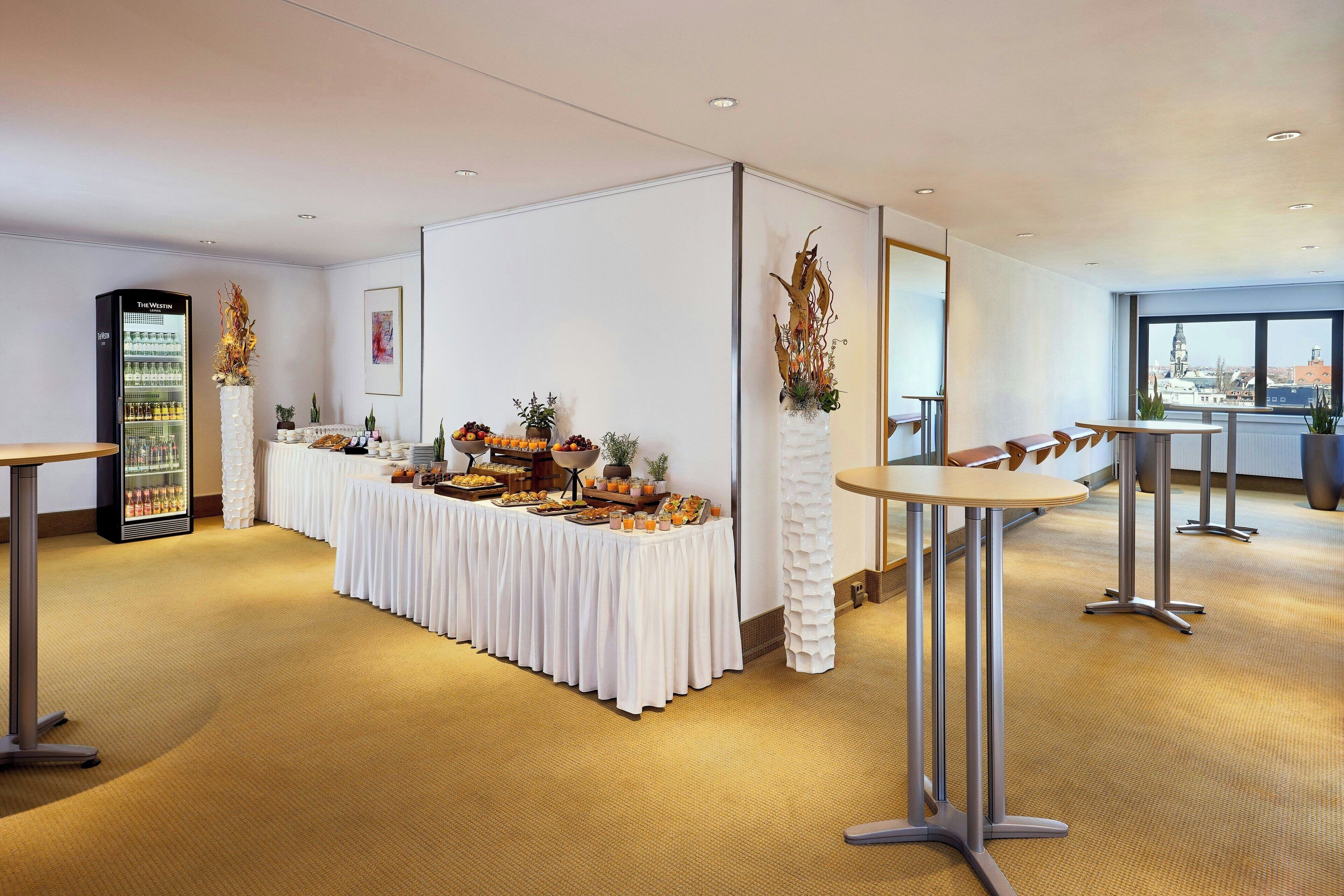 A modern hotel room with a buffet setup, a minibar, and a view of the city through large windows.