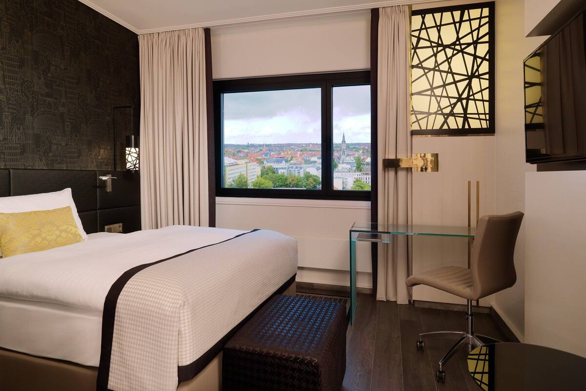 Habitación de hotel con una cama grande, un escritorio, una silla y vistas a la ciudad a través de una ventana.