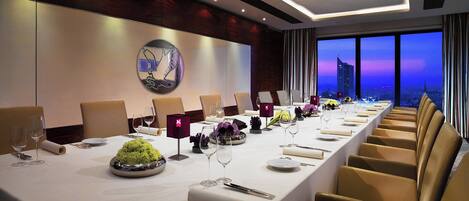 A long dining table with multiple chairs, set for a formal meal, in a modern hotel room with a city view.