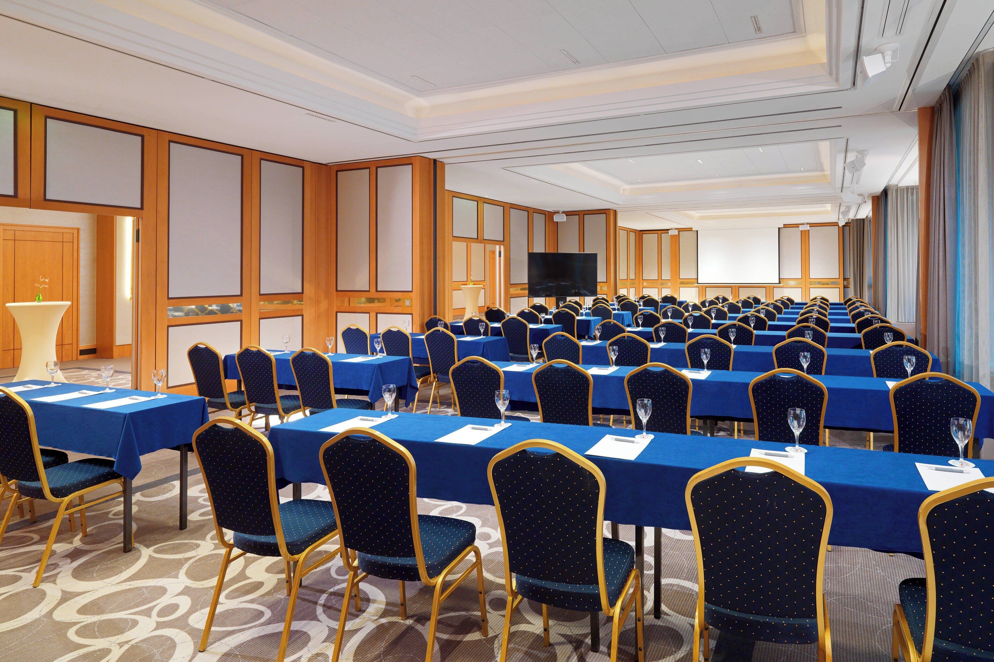 Un salón de conferencias con filas de mesas y sillas, una pantalla grande y un proyector.