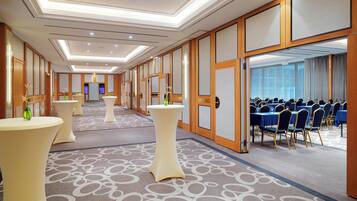 Un vestíbulo de hotel con un mostrador de recepción alargado, varias mesas redondas y una sala de conferencias con mesas y sillas azules.