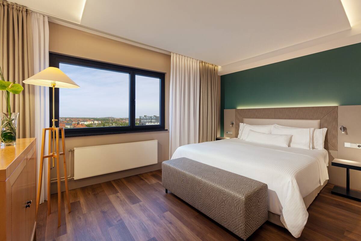 Habitación de hotel moderna con una cama grande, un escritorio con lámpara, vistas de la ciudad a través de un ventanal amplio y un radiador debajo de la ventana.