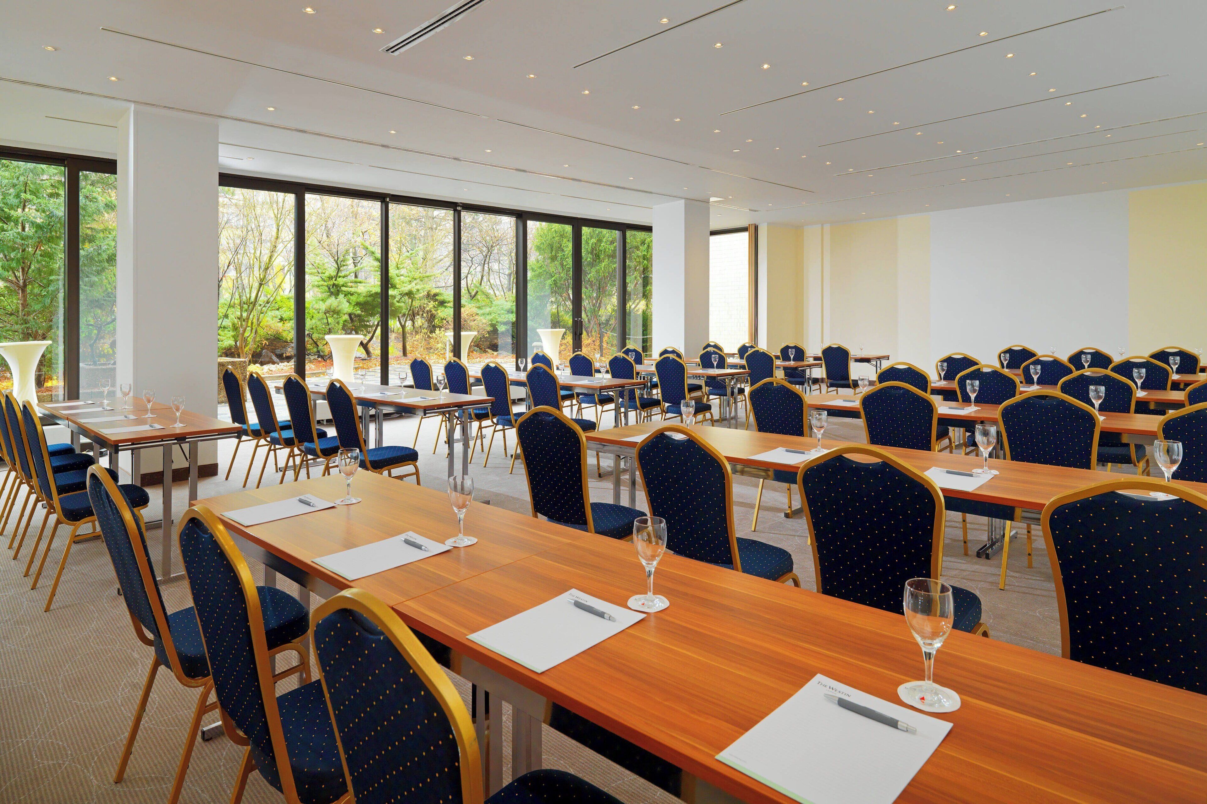 A conference room with multiple tables and chairs, large windows with a view of trees, and a carpeted floor.