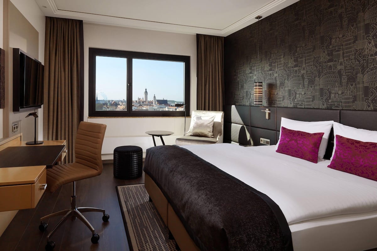 Habitación de hotel con una cama grande, un escritorio, una silla y vistas a un paisaje urbano a través de una ventana.