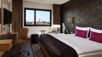 Ein Hotelzimmer mit einem großen Bett, einem Schreibtisch, einem Sessel und Blick auf eine Stadtlandschaft durch ein Fenster.