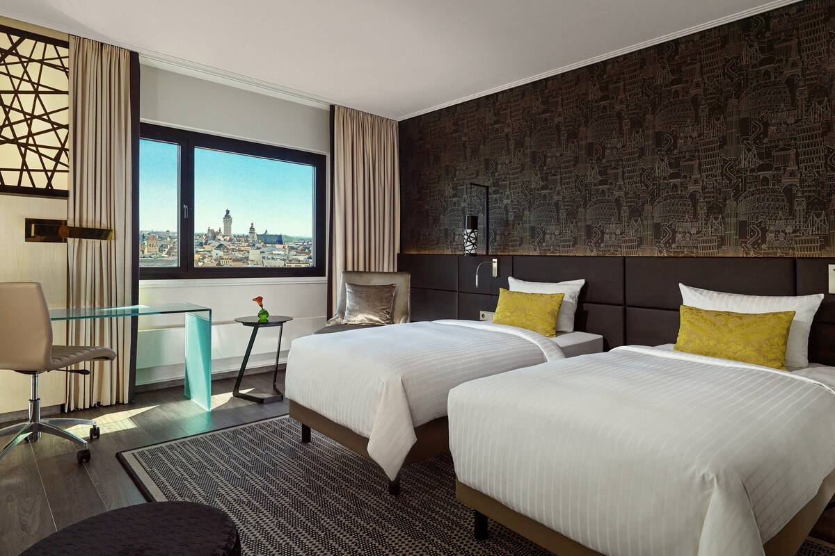 Habitación de hotel con dos camas, un escritorio de vidrio, vistas a la ciudad y una pared con diseño.