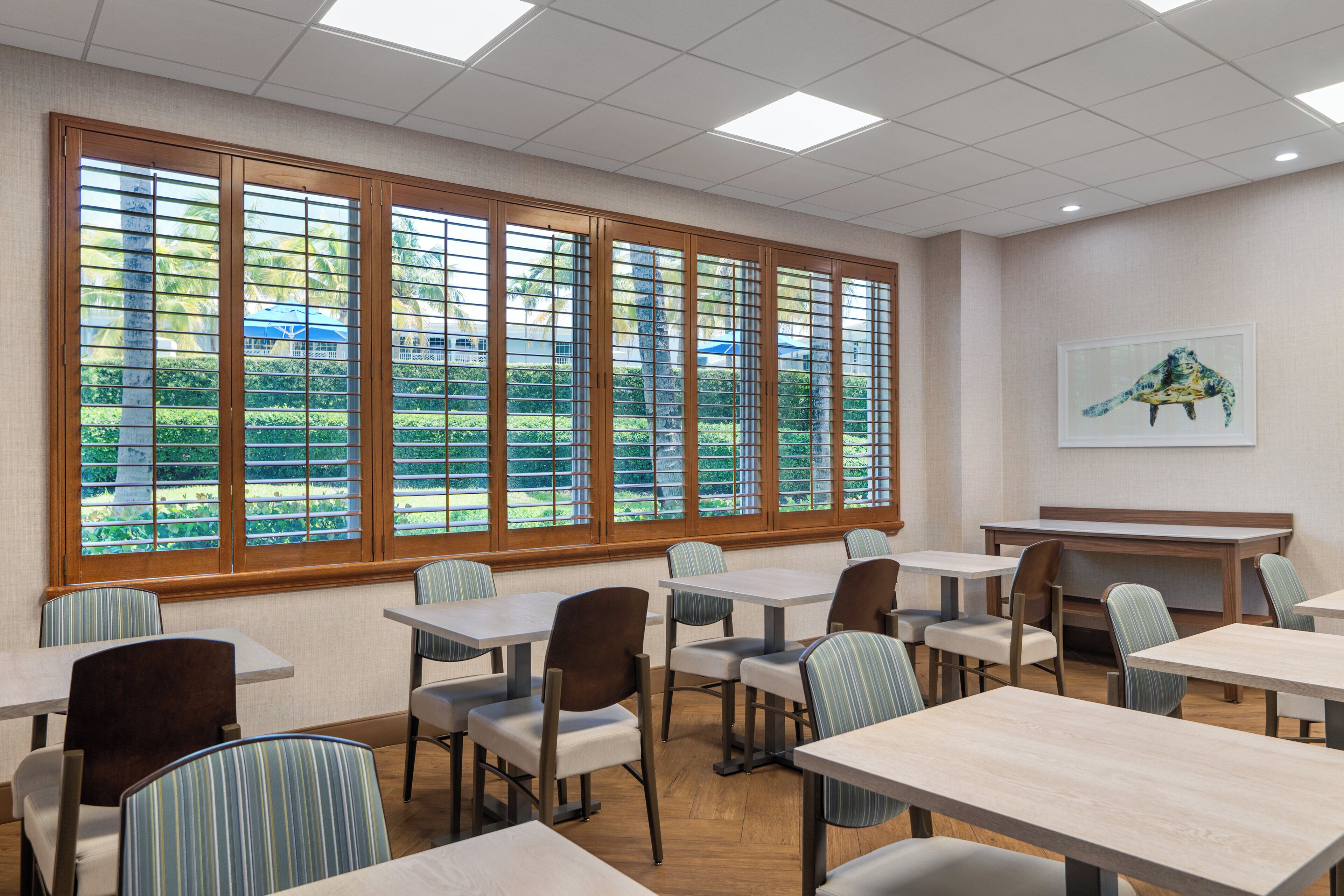 A dining area with tables and chairs, large windows with blinds, and a painting on the wall.
