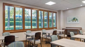 A dining area with tables and chairs, large windows with blinds, and a painting on the wall.