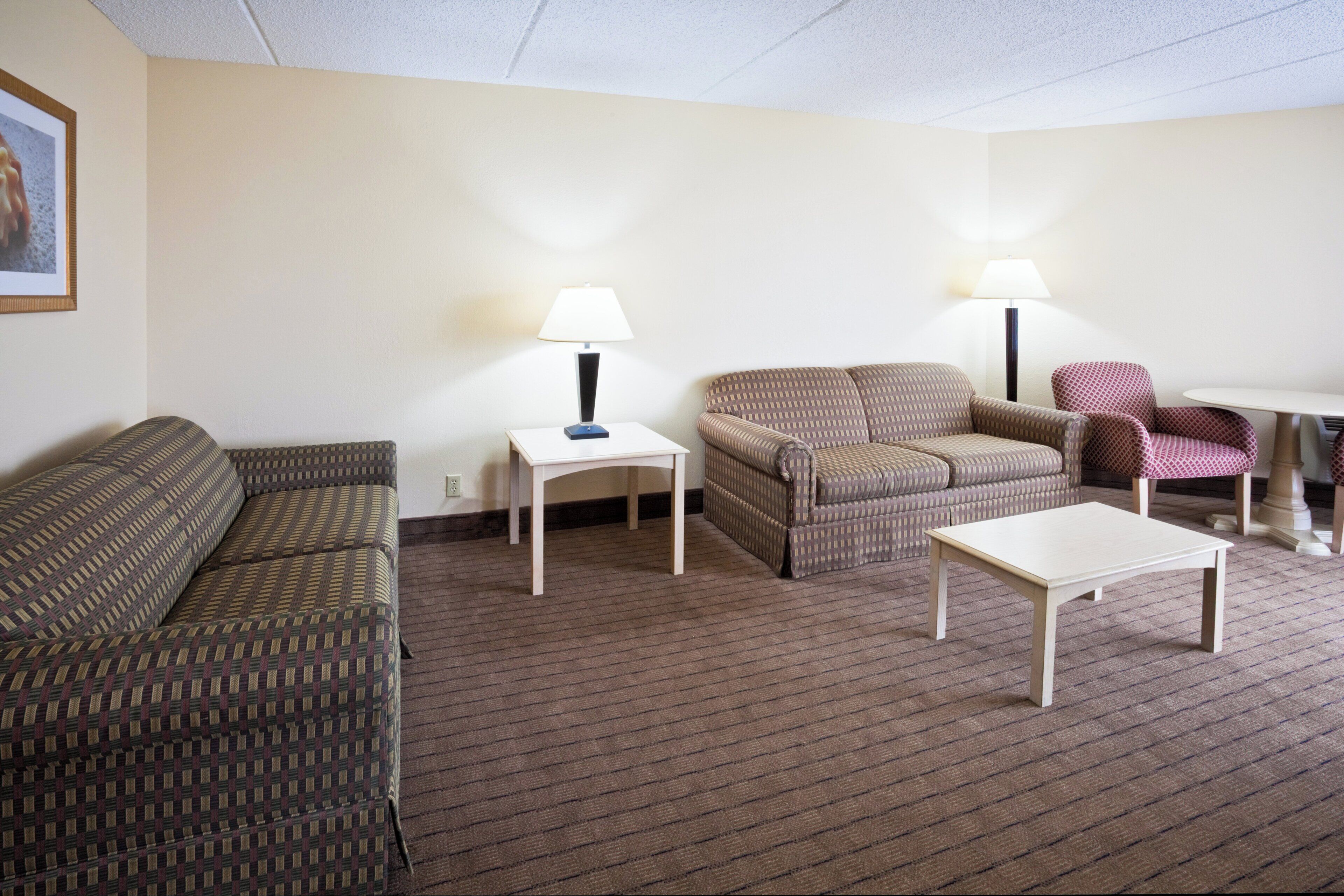 A hotel room with a patterned sofa, a small white table, a patterned armchair, and a lamp on a side table.