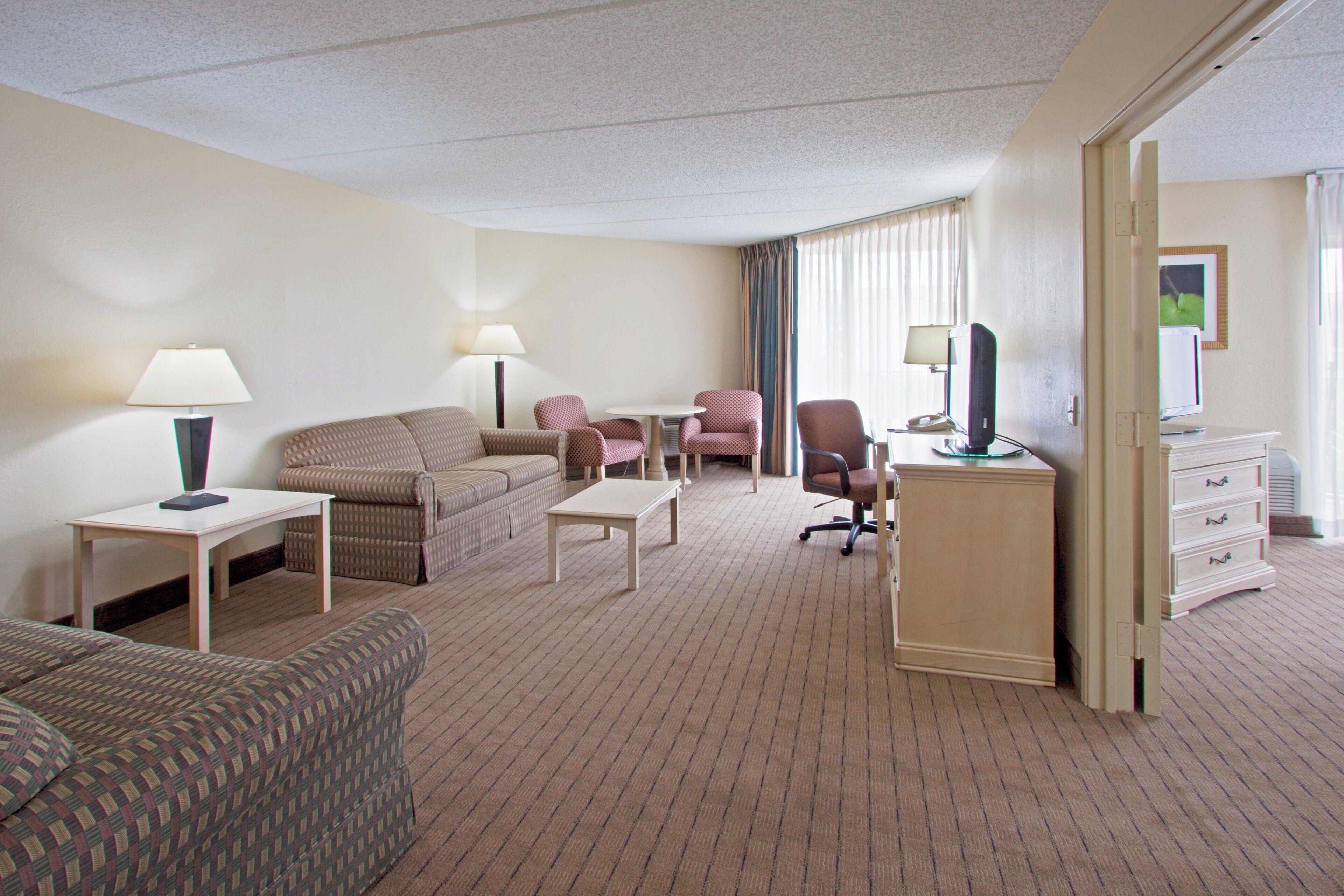 Une chambre d’hôtel comprenant un canapé, deux fauteuils, une petite table, un bureau avec un ordinateur, une commode et une lampe.