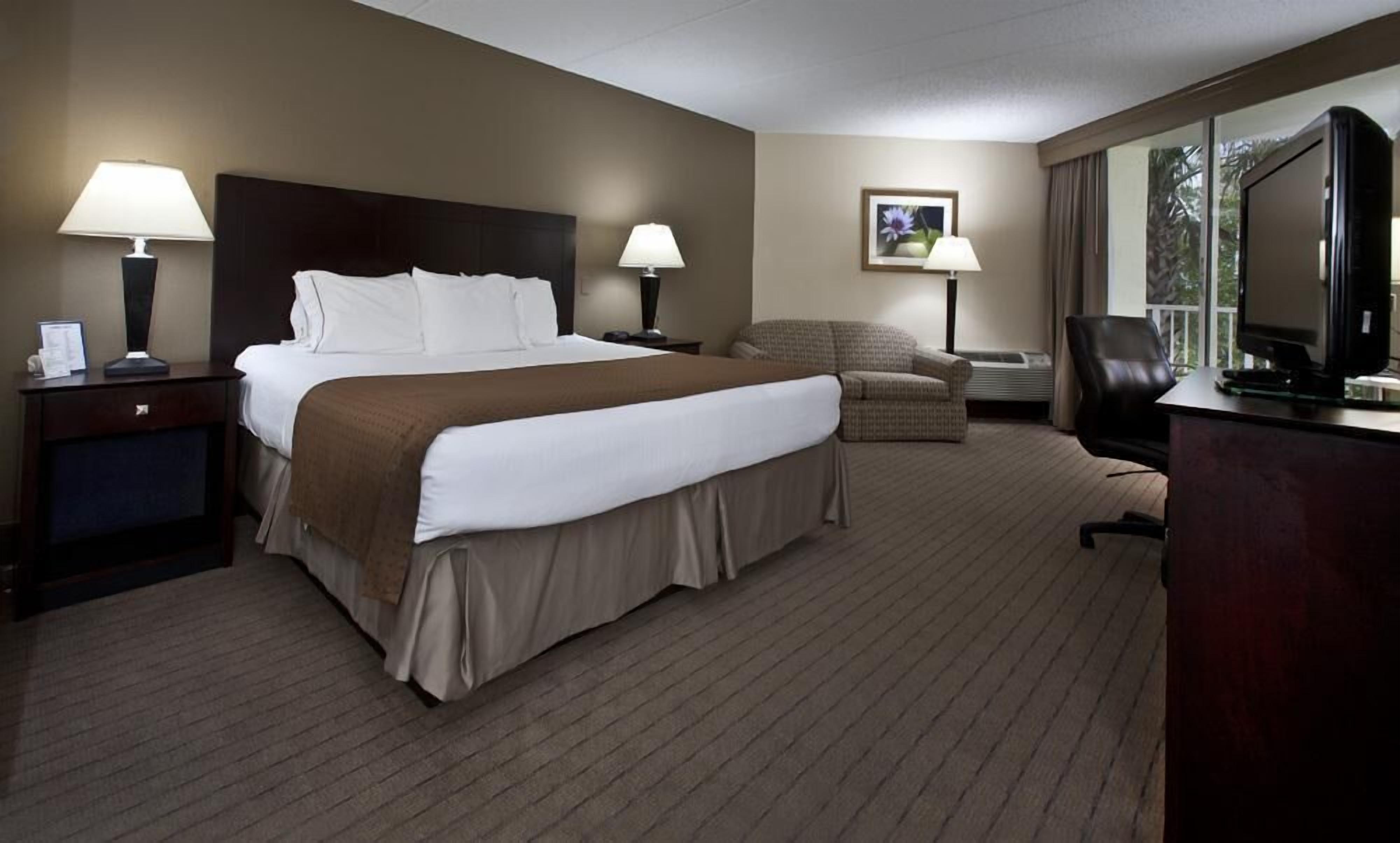 A hotel room with a large bed, two bedside tables with lamps, a chair, a desk with a computer, and a television.