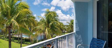 A balcony with a blue chair, a white railing, and a view of palm trees and a pool.