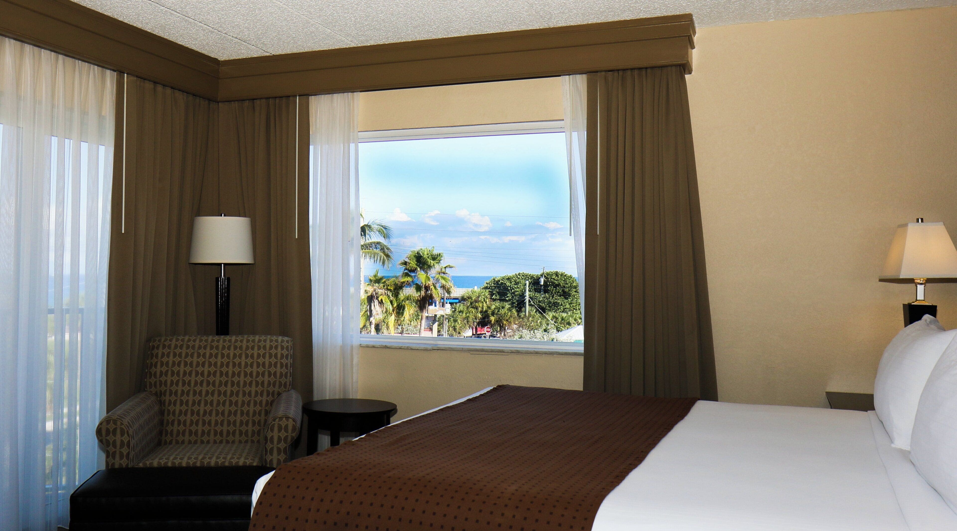 Habitación de hotel con cama, una silla, una mesita y ventana que ofrece vista a palmeras y un edificio.