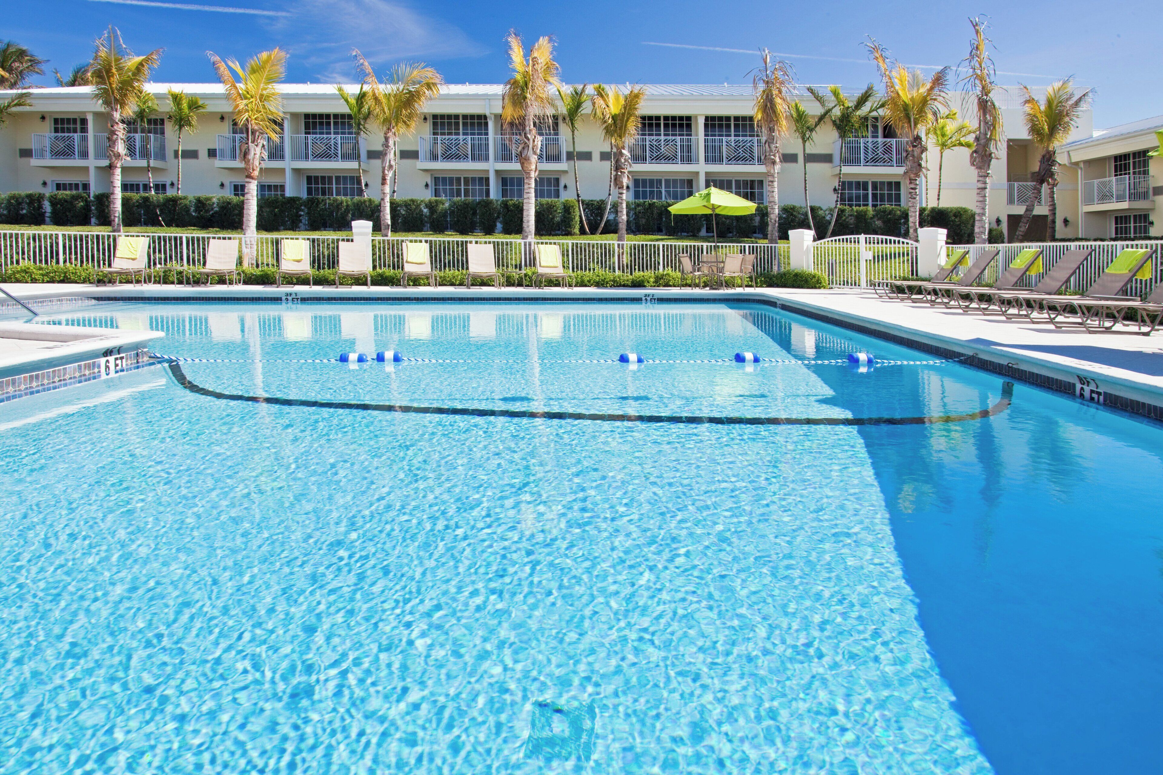 Une piscine entourée de chaises longues et de palmiers, le tout bordé d’un immeuble doté de balcons.