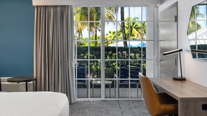 Une chambre d’hôtel comprenant un lit, un bureau, une chaise, une petite table et une vue sur les palmiers, visible à travers une porte-fenêtre coulissante.