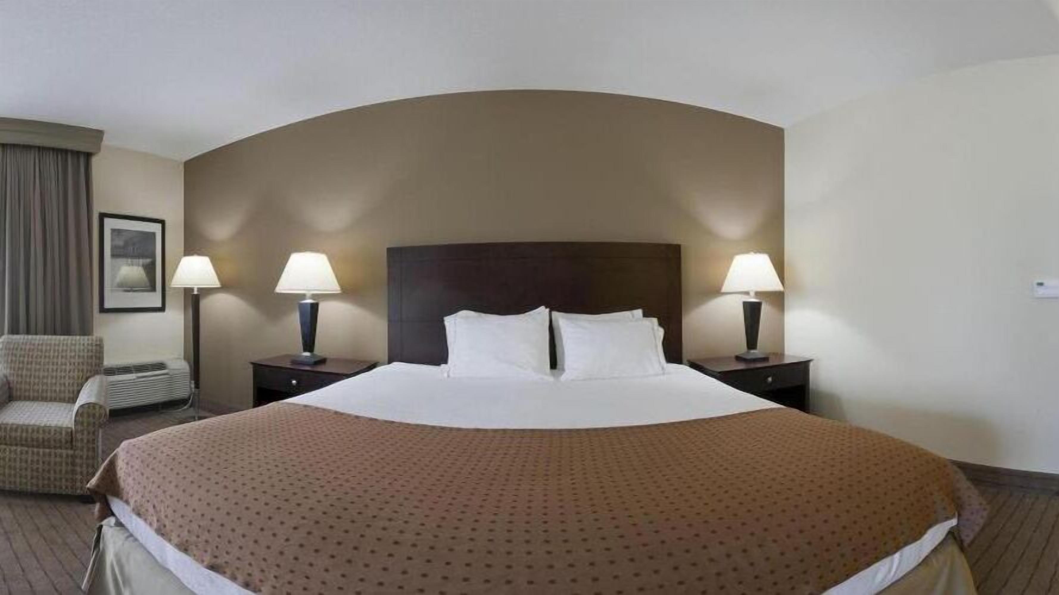 A hotel room with a large bed, two bedside lamps, a chair, and a framed picture on the wall.