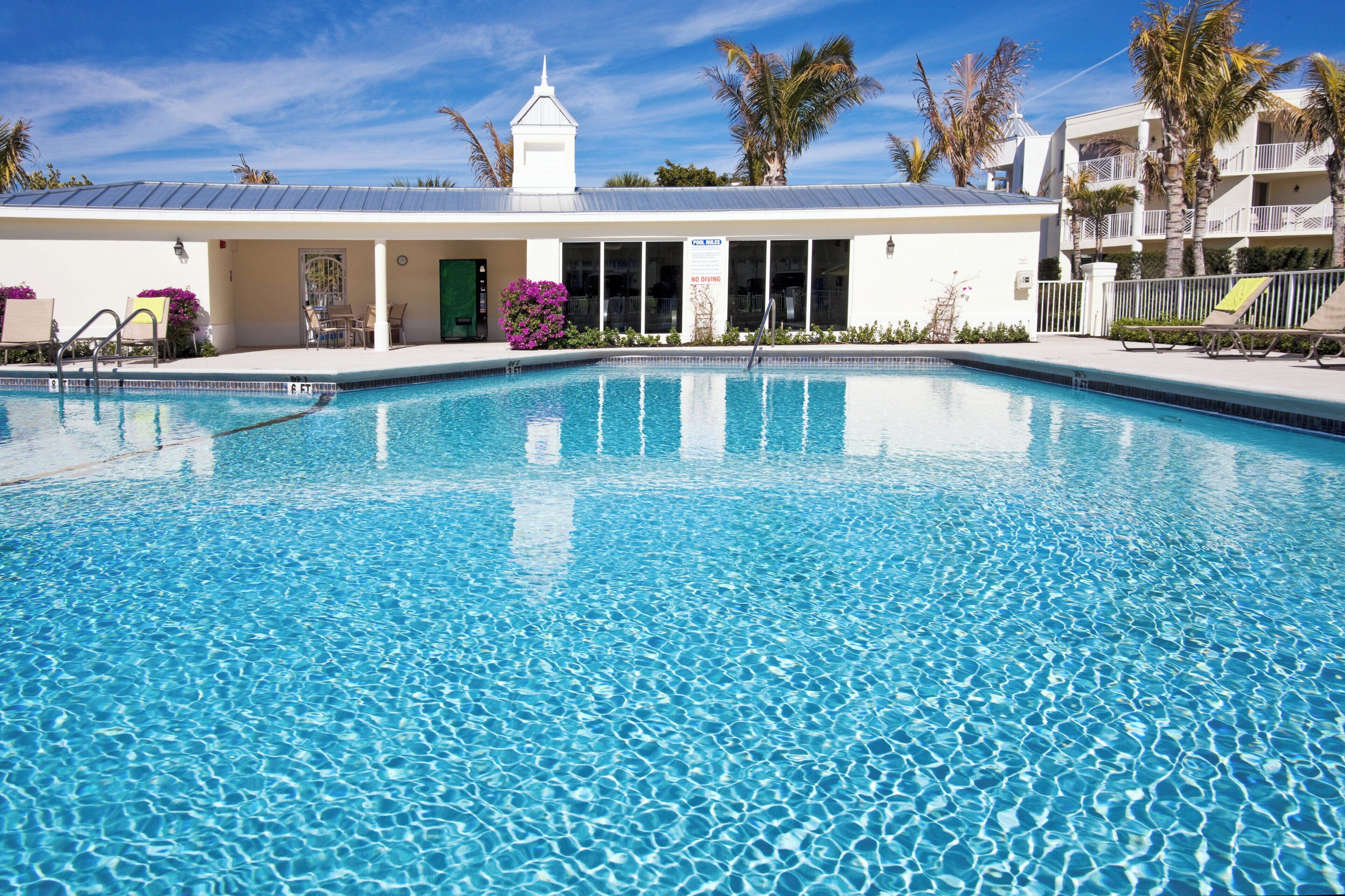 Piscina límpida com posto de salva-vidas, área de estar coberta e edifício com fachada branca e um relógio.