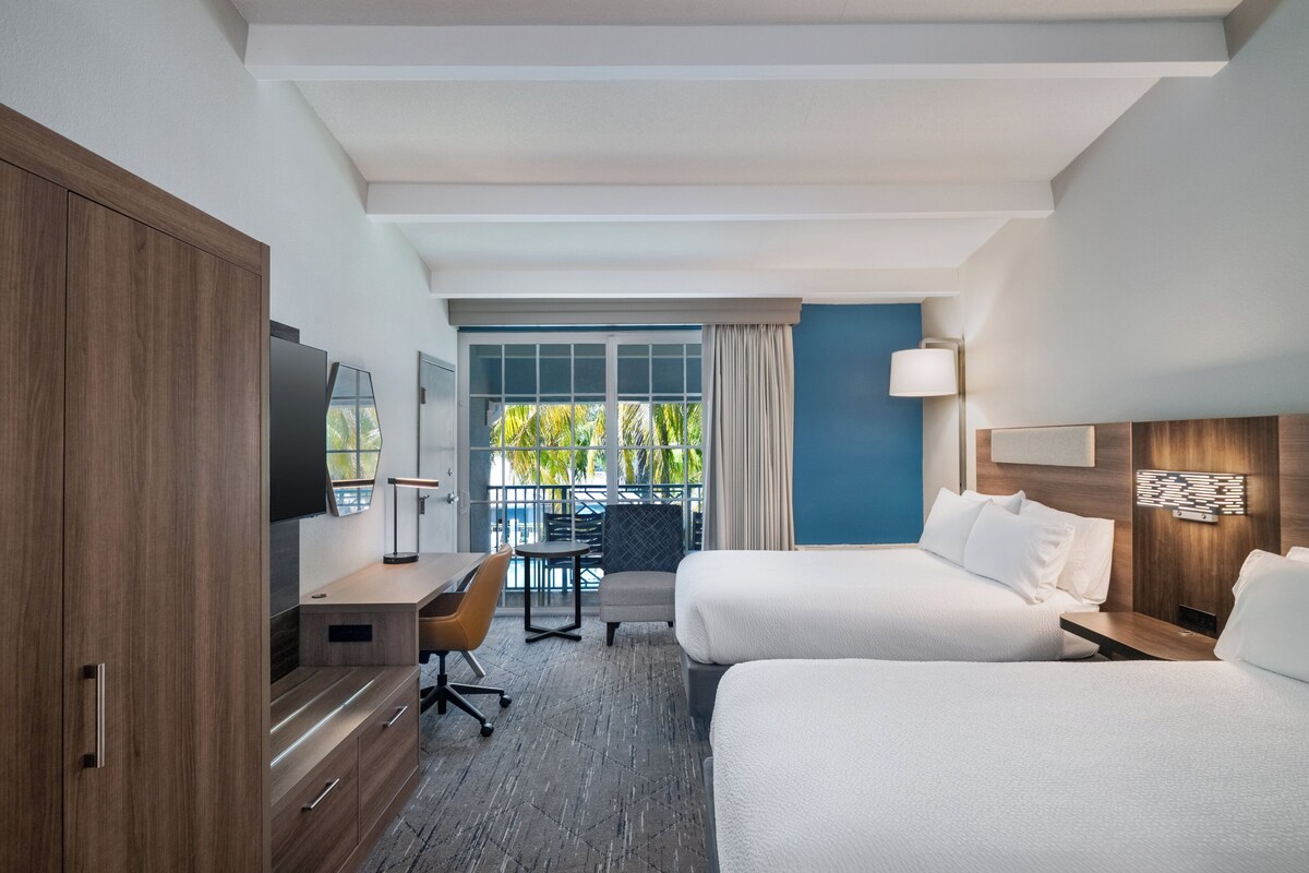 Une chambre d’hôtel avec deux lits, un bureau, une télévision et un balcon offrant une vue sur les palmiers.