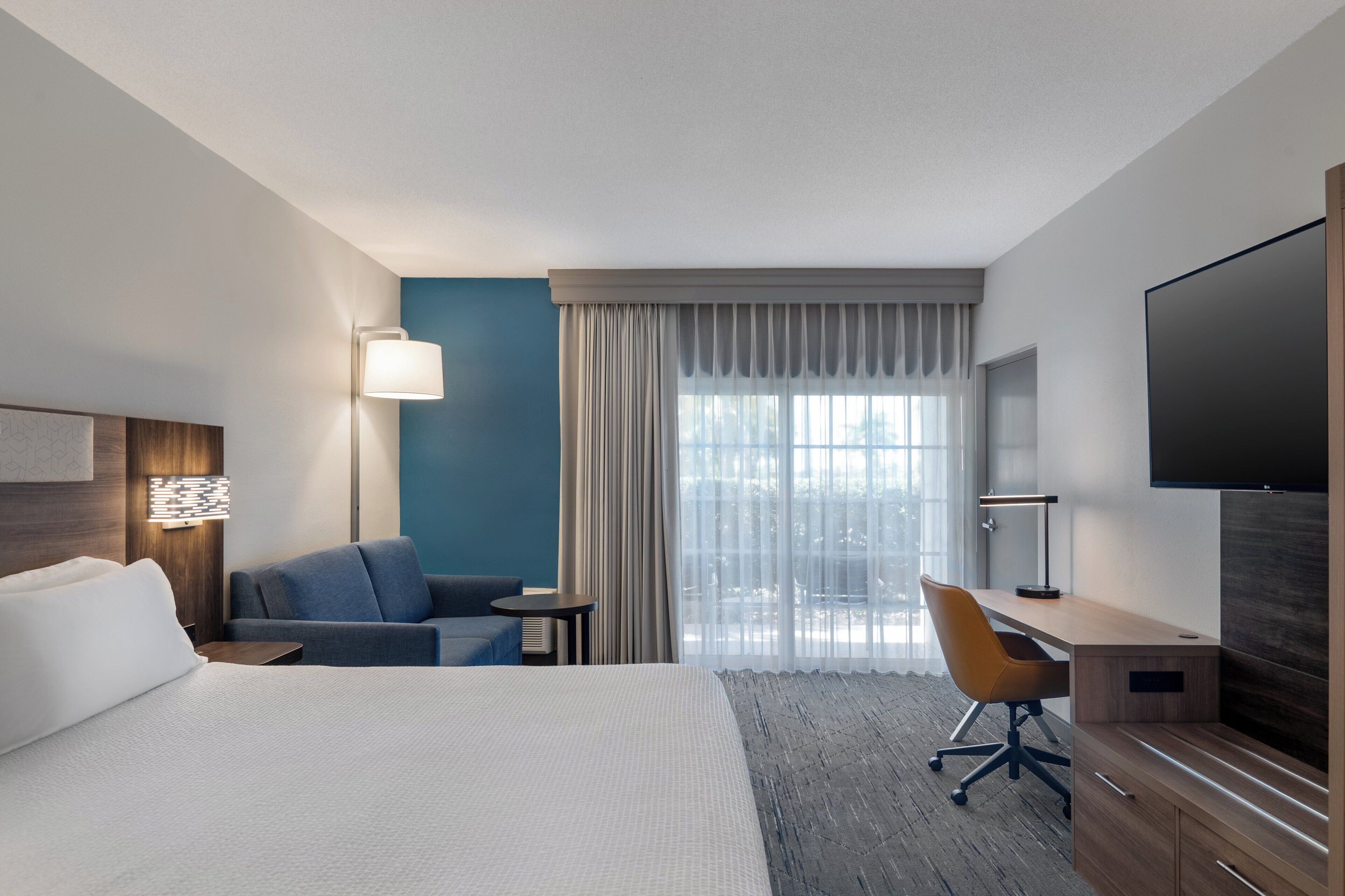 Habitación de hotel con cama, escritorio, silla, televisión, lámpara y vistas al exterior.
