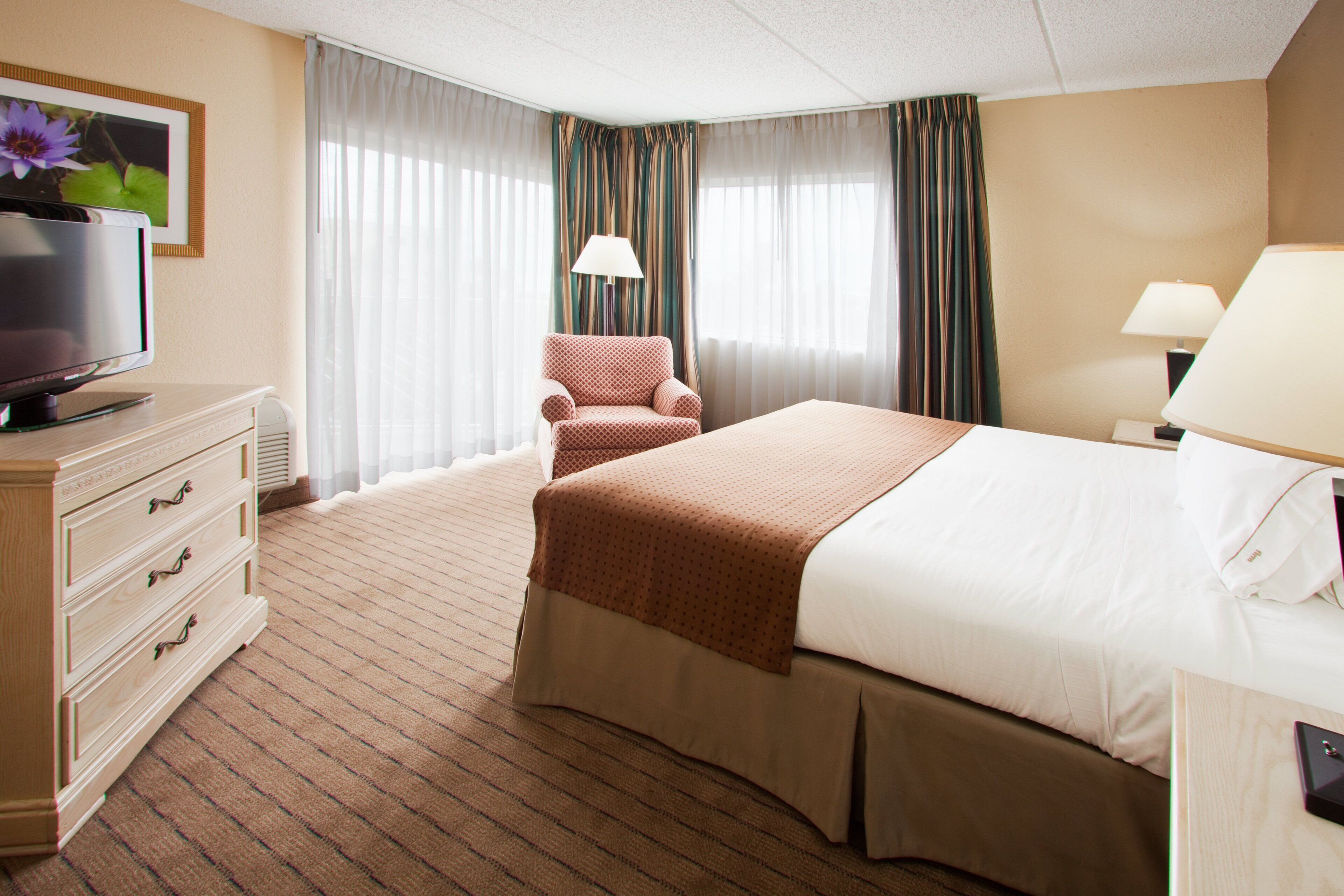 A hotel room with a bed, a television on a dresser, a chair, and a lamp.