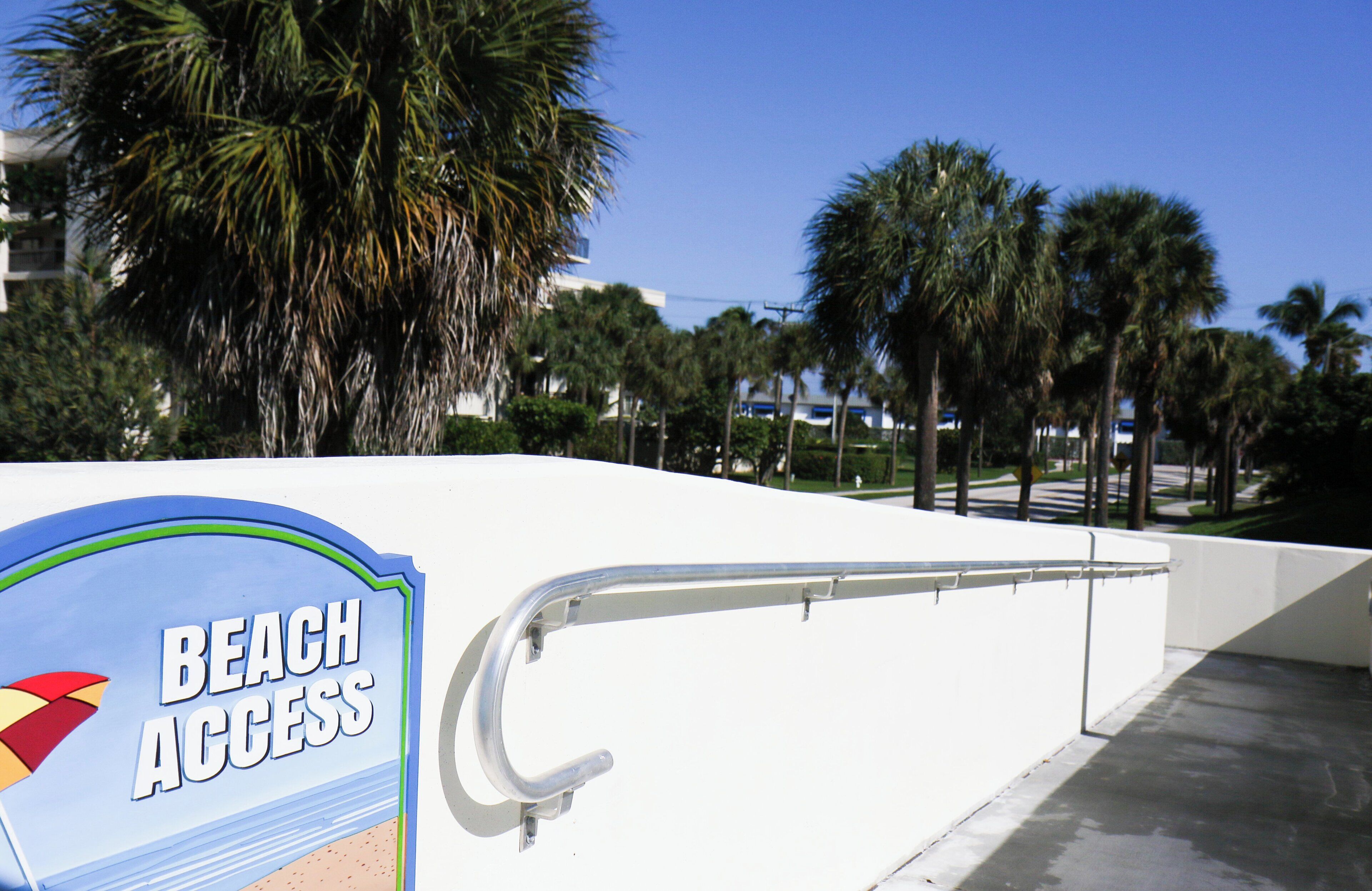 A sign indicating beach access, palm trees, and a white building in the background.