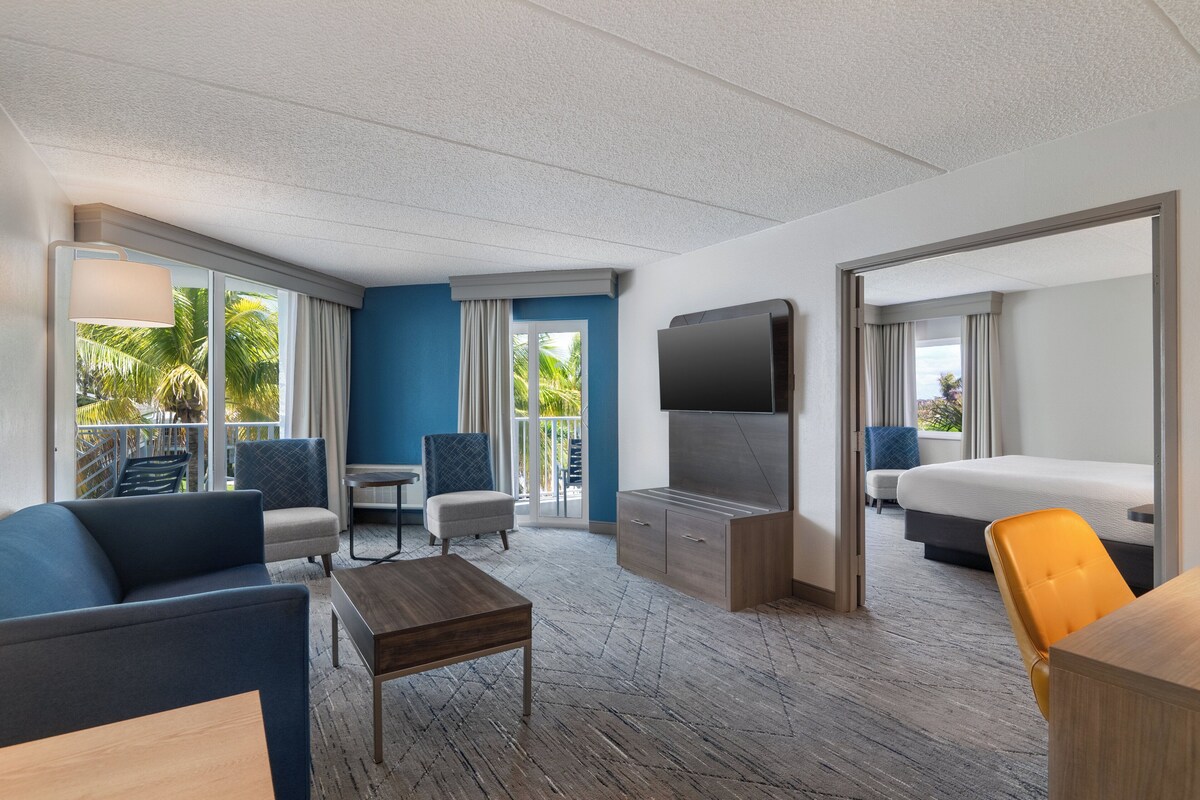 Une chambre d’hôtel avec un grand lit, une télévision, un canapé, une table basse et un balcon offrant une vue sur les palmiers.