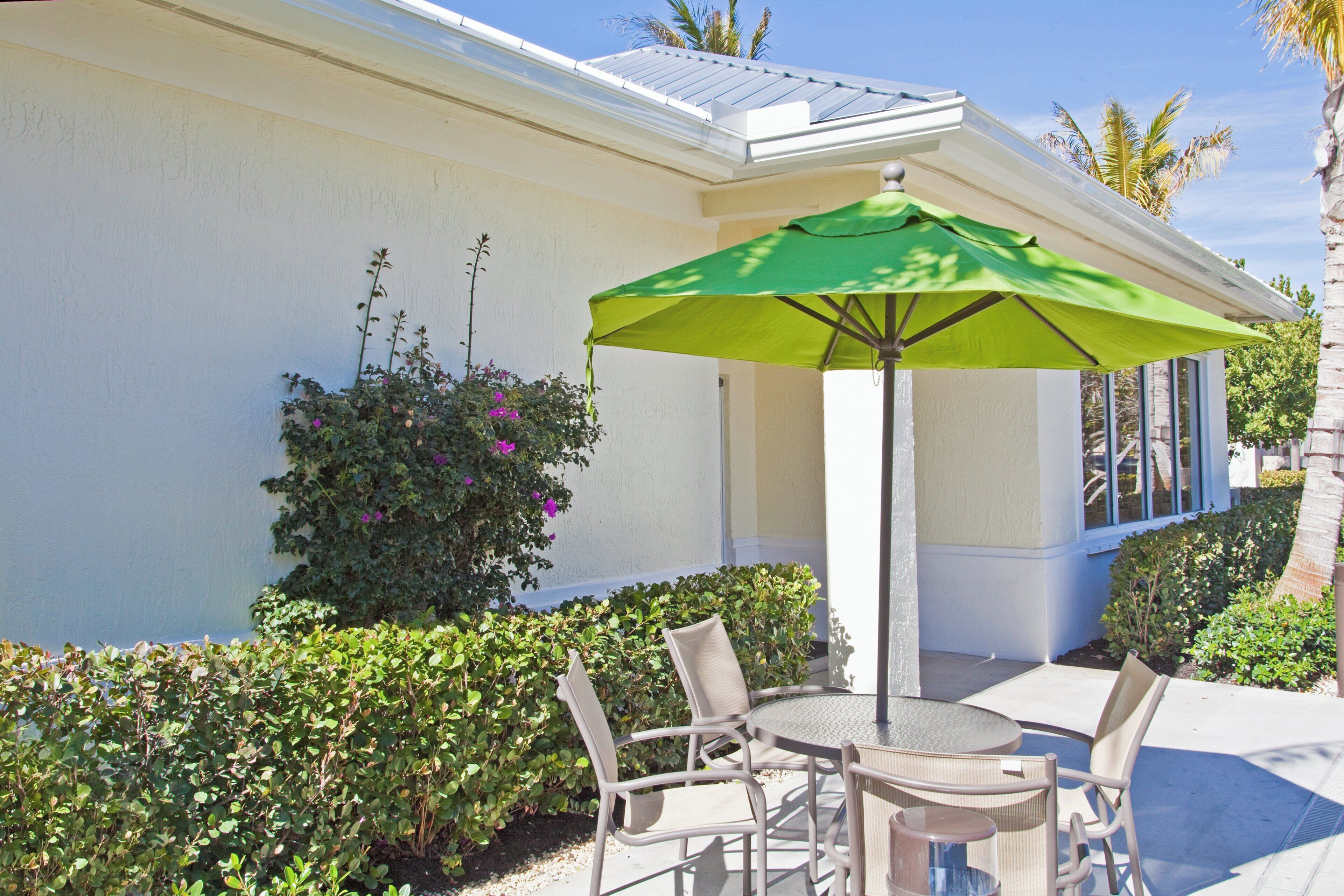 An outdoor patio with a table and chairs, a green umbrella, and a white building with a window.