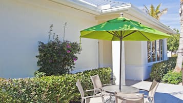 An outdoor patio with a table and chairs, a green umbrella, and a white building with a window.
