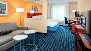 Ein Hotelzimmer mit einer Couch, zwei kleinen Tischen, einem Bett mit blauer Bettdecke, einem Schreibtisch mit Computer und einem Fernseher.