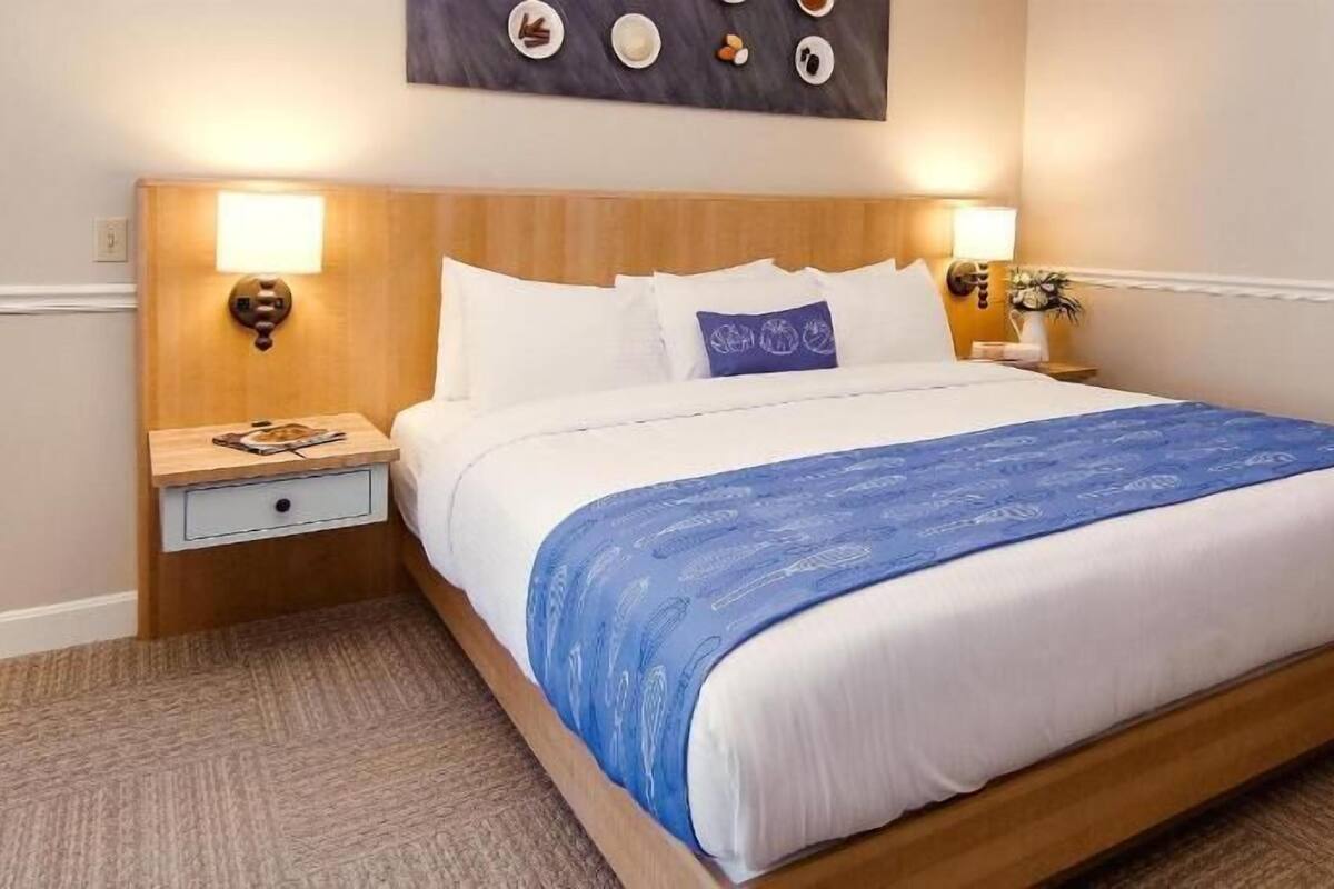 A hotel room with a large bed, wooden headboard, two bedside tables with lamps, a small nightstand, and a wall-mounted artwork.