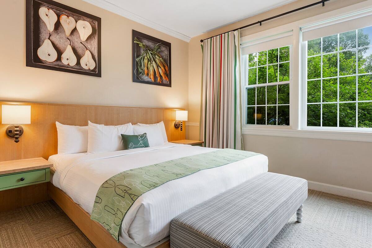 A hotel room with a large bed, two bedside tables, a bench, a window with curtains, and artwork on the walls.