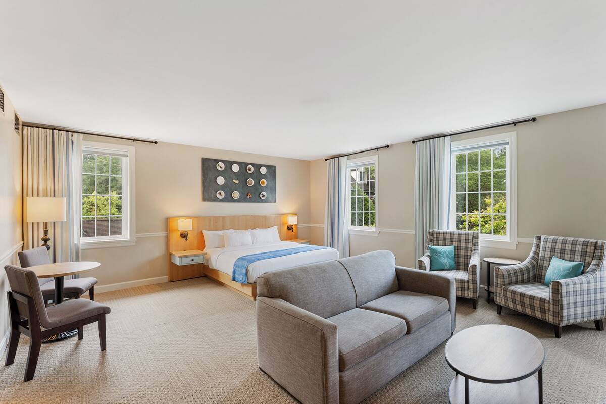 A hotel room with a bed, a sofa, a chair, a small table, and a lamp.