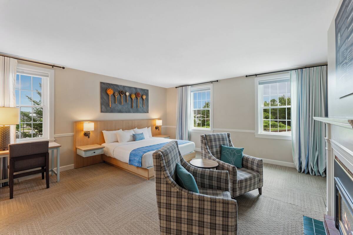 A hotel room with a bed, two armchairs, a desk, a lamp, a painting, and a fireplace.
