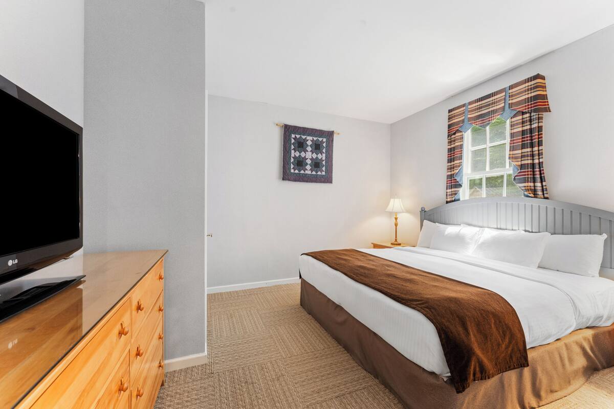 A hotel room with a bed, a TV, a wooden dresser, a wall-mounted quilt, and a window with curtains.