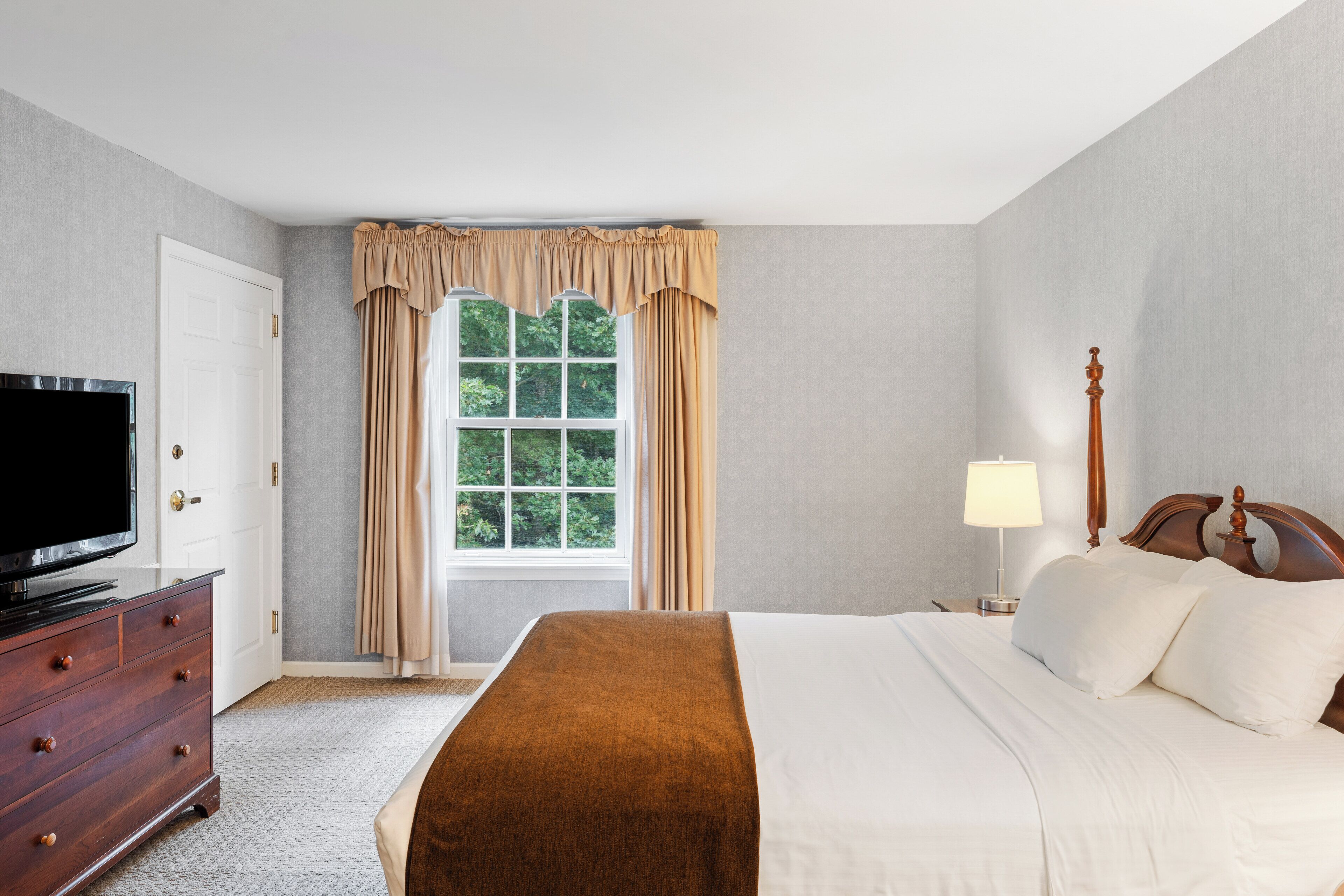 Habitación de hotel con cama, televisión, cómoda, ventana con cortinas y mesita de noche con lámpara.
