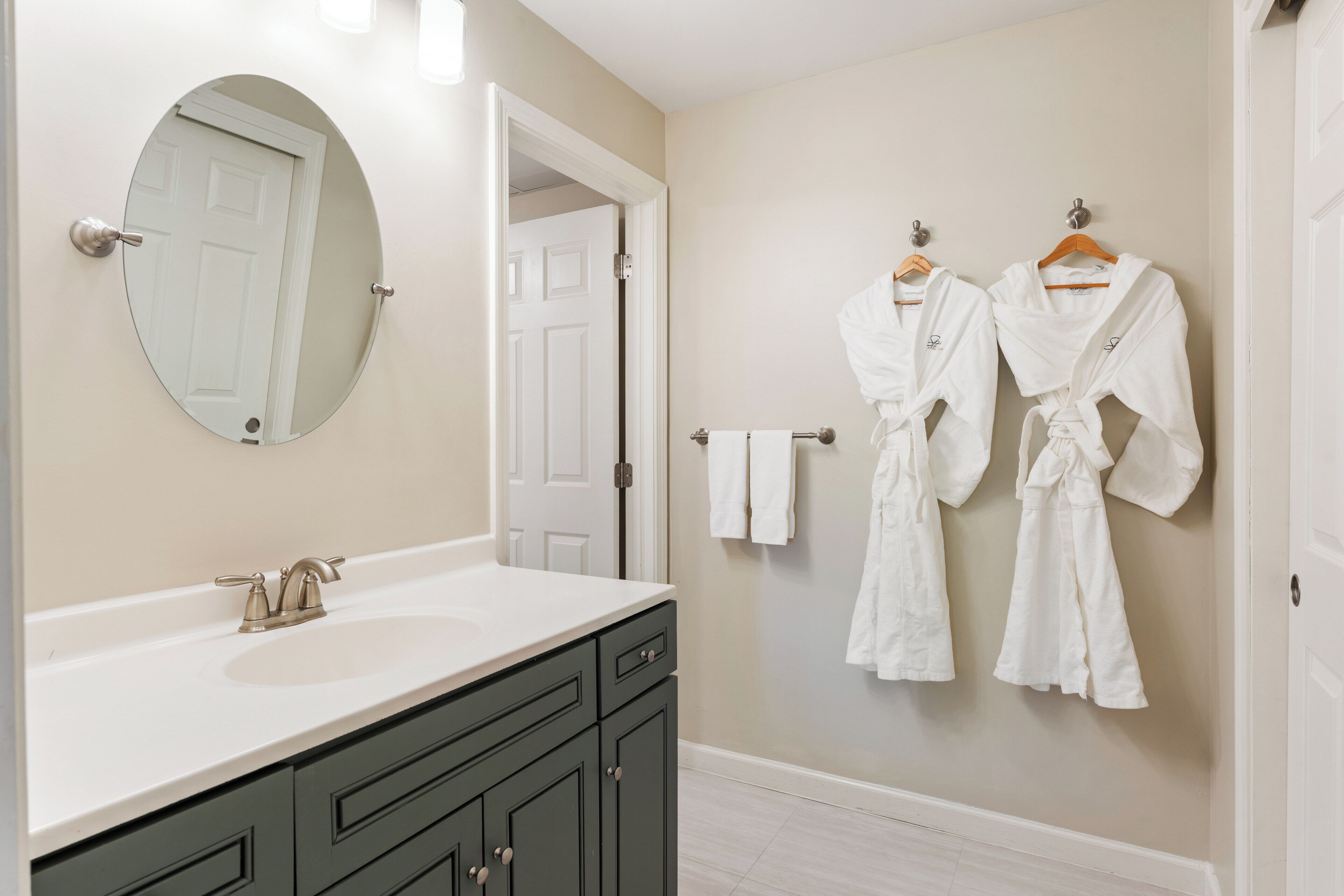 Un baño con un lavabo blanco, muebles oscuros, un espejo redondo y dos albornoces blancos colgados en la pared.