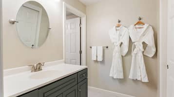 A bathroom with a white sink, dark cabinets, a round mirror, and two white robes hanging on the wall.