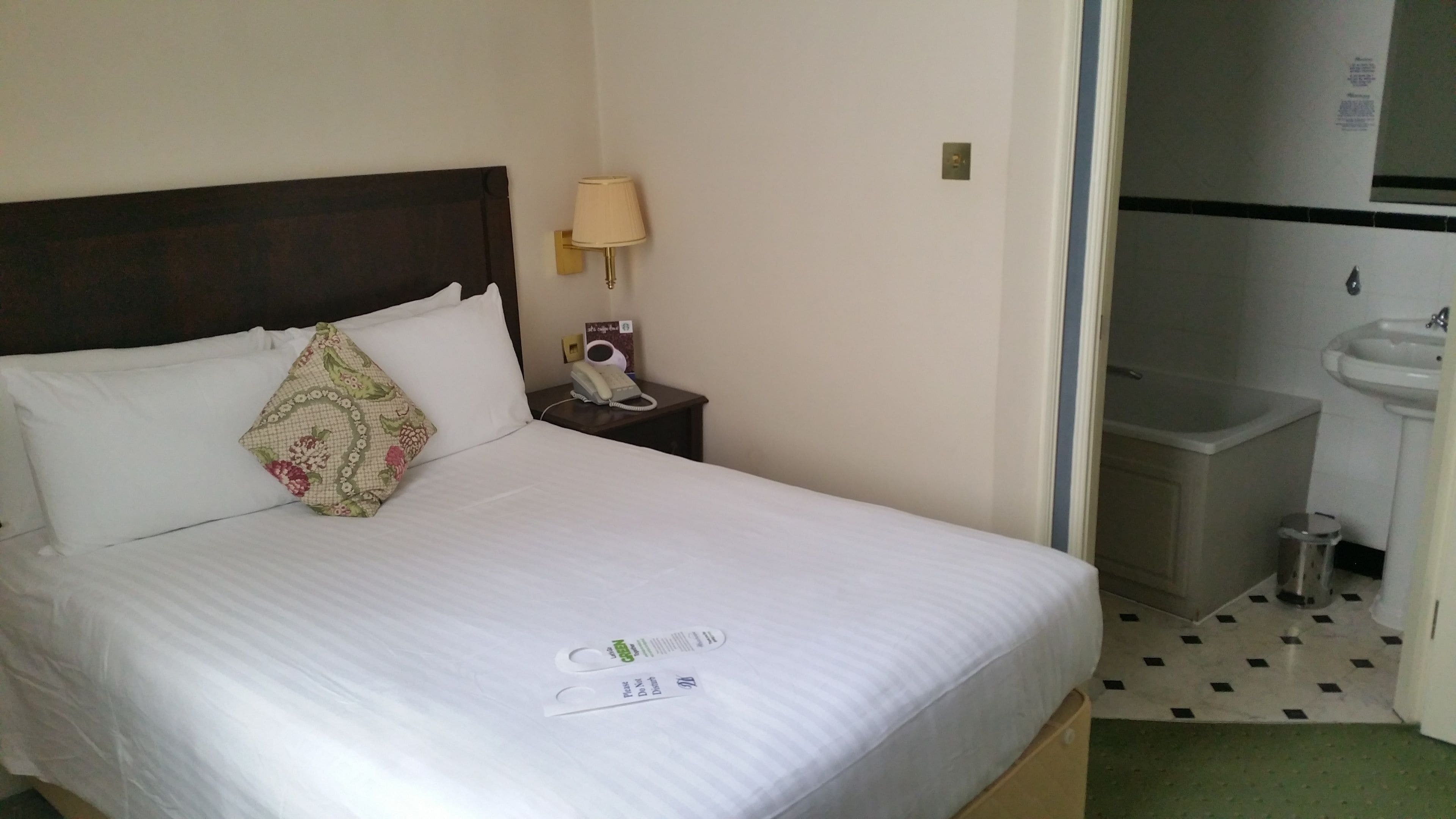A hotel room with a bed, a bedside table with a lamp, a telephone, and a bathroom with a sink and toilet.
