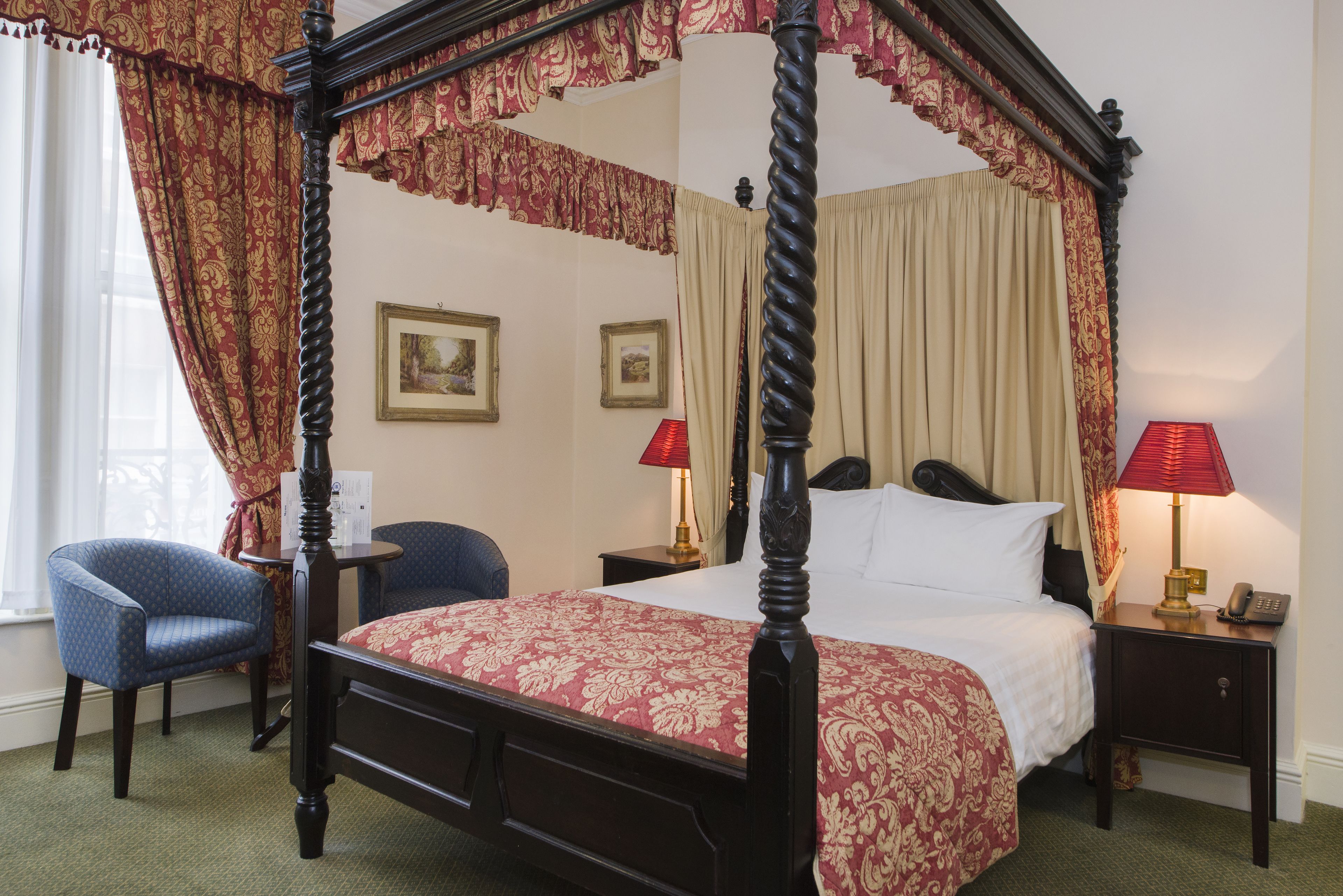 A four-poster bed with a red and gold bedspread, two bedside tables with lamps, a chair, and framed pictures on the wall.